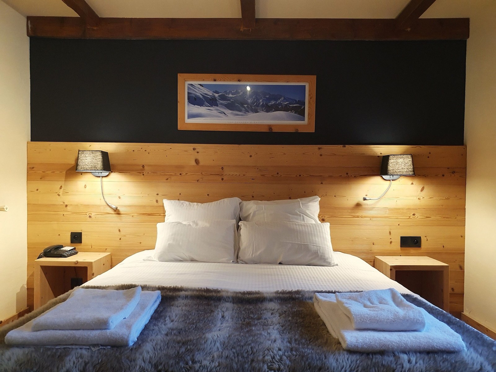 A bedroom with a wooden headboard, two bedside tables, a bed with white bedding, a mounted picture, and two wall-mounted lamps. - Chalet Hotel Du Bois (Les Houches)