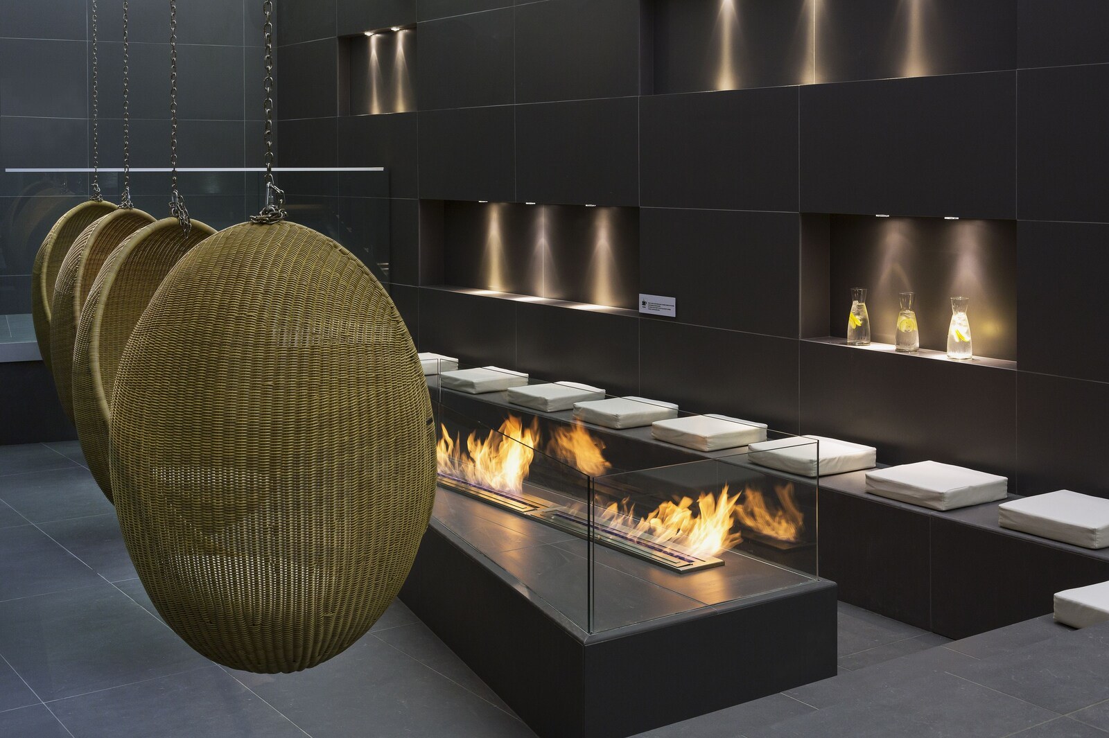 A modern interior with hanging wicker chairs, a glass-top table with a built-in fire feature, and shelves with bottles. - Héliopic Hôtel & Spa (Chamonix-Mont-Blanc)