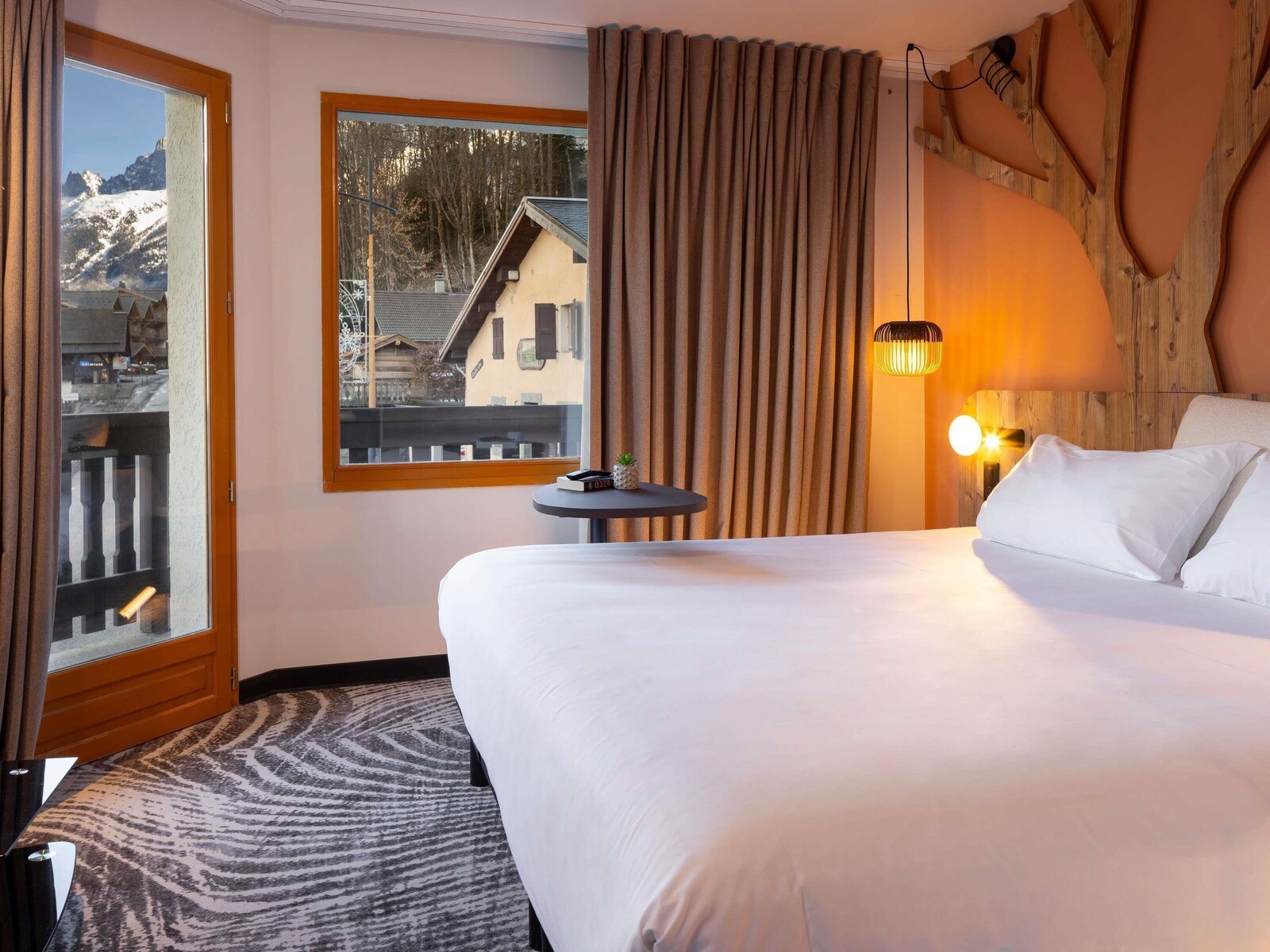 A hotel room with a large bed, a small table, a window with a view of a mountain, and a wooden wall. - Ibis Styles Les Houches Chamonix (Les Houches)