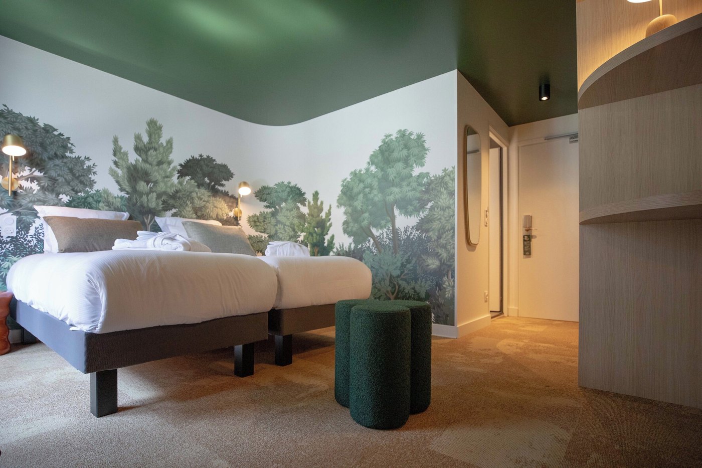 A bedroom with two separate beds, pillows, two green ottomans, and a wall mural of trees. — Lykke Chamonix