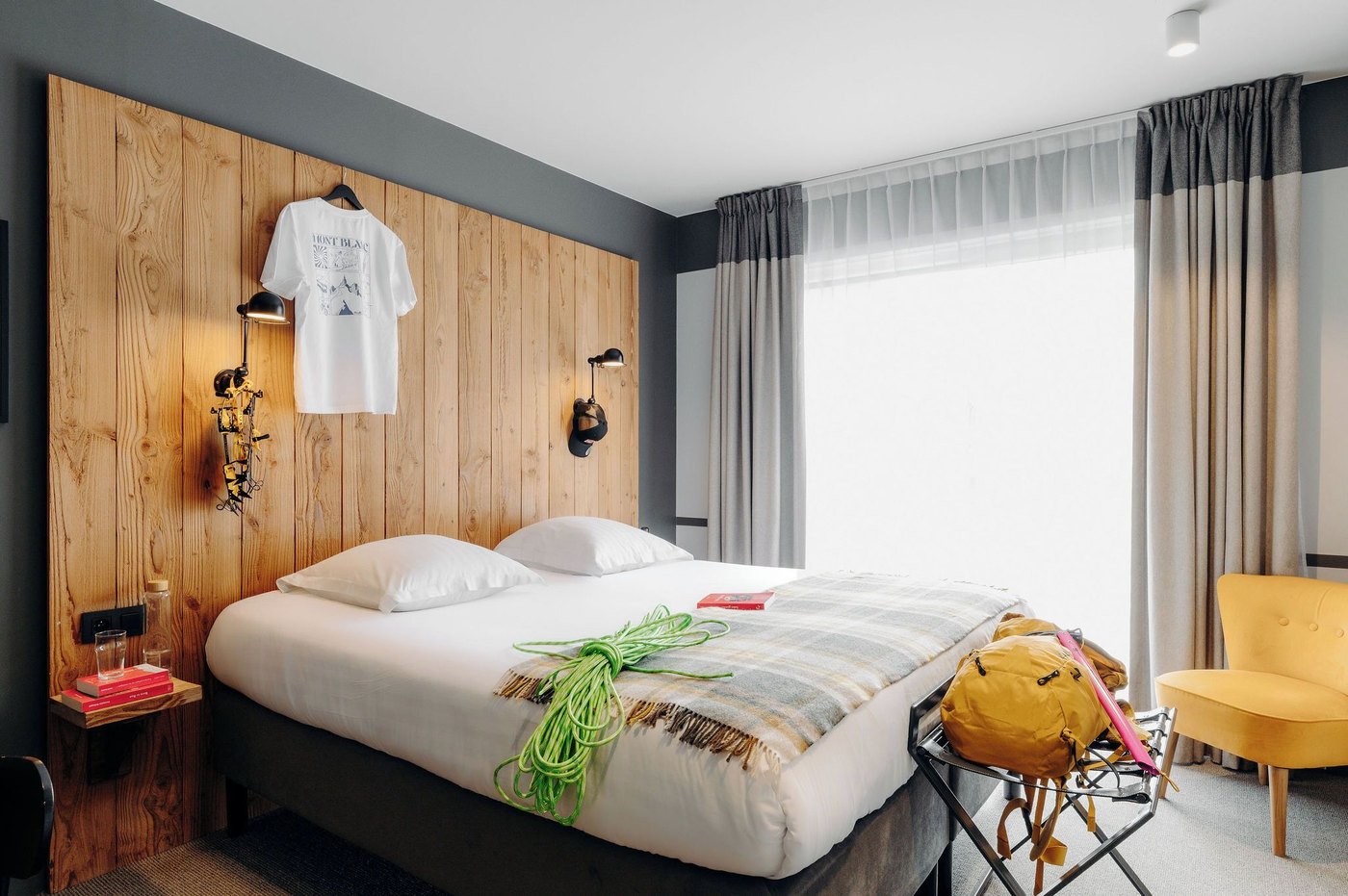 Modern PlanB Living Chamonix guest room with a wooden headboard, white bed linen, yellow chair and large curtained window.